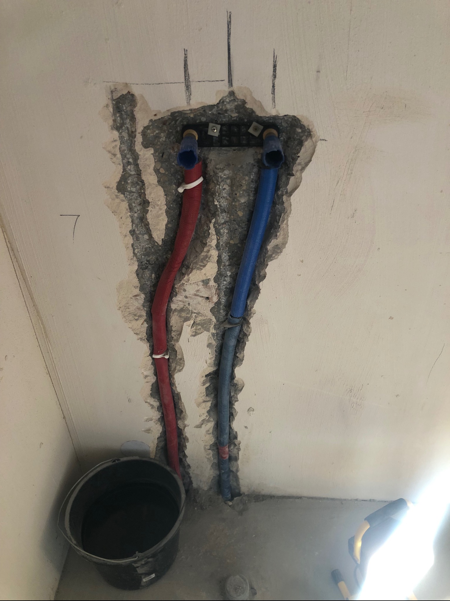 Fragment ściany z instalacją wodną: czerwona i niebieska rura prowadzą do metalowej rozdzielnicy. Widoczne ślady po kuciu w ścianie i wiadro na podłodze.