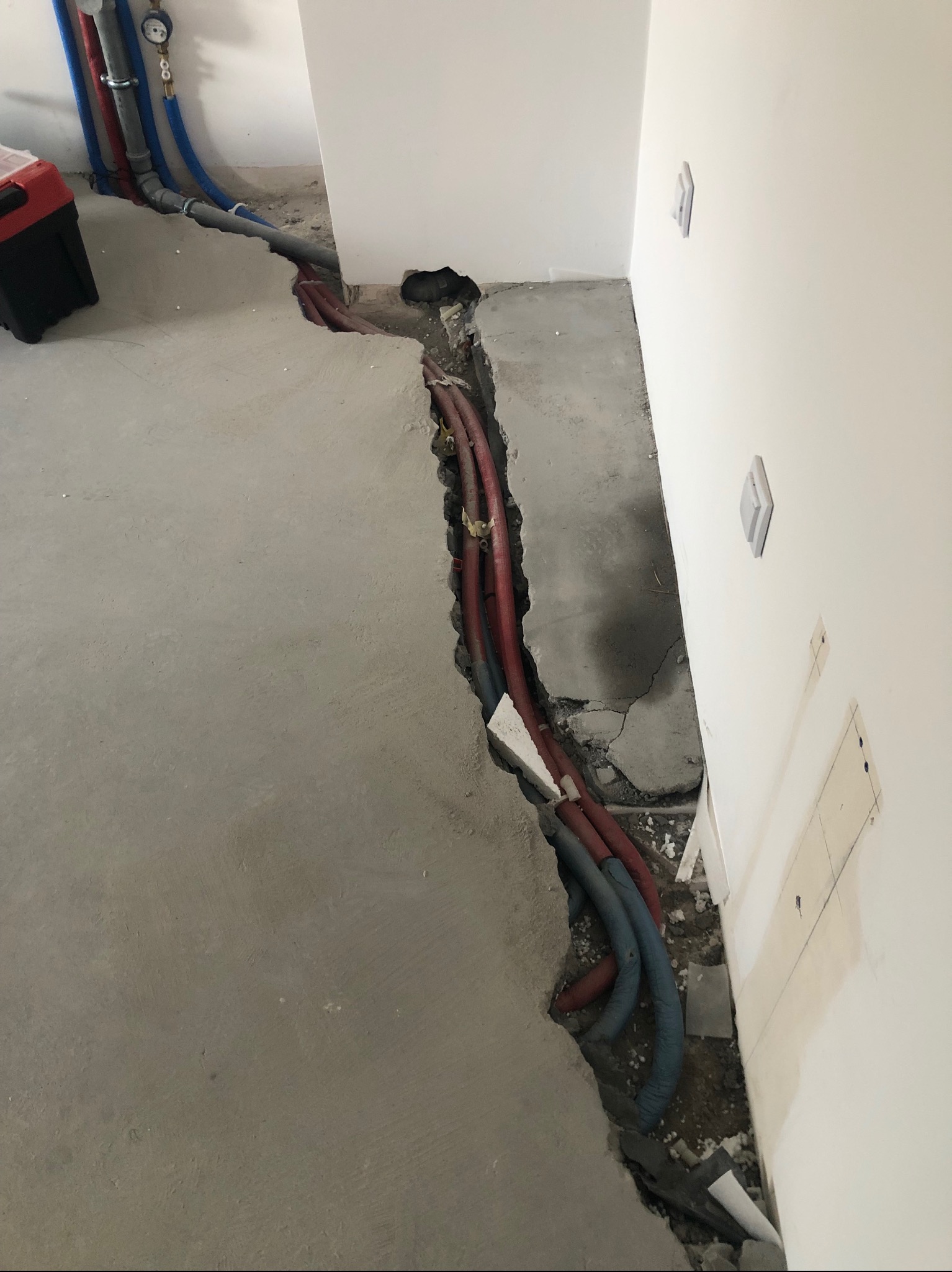 Wykop w szarej posadzce z widocznymi rurami instalacji wodnej i grzewczej, częściowo zakrytymi. Na ścianie widoczne puszki elektryczne i włączniki.
