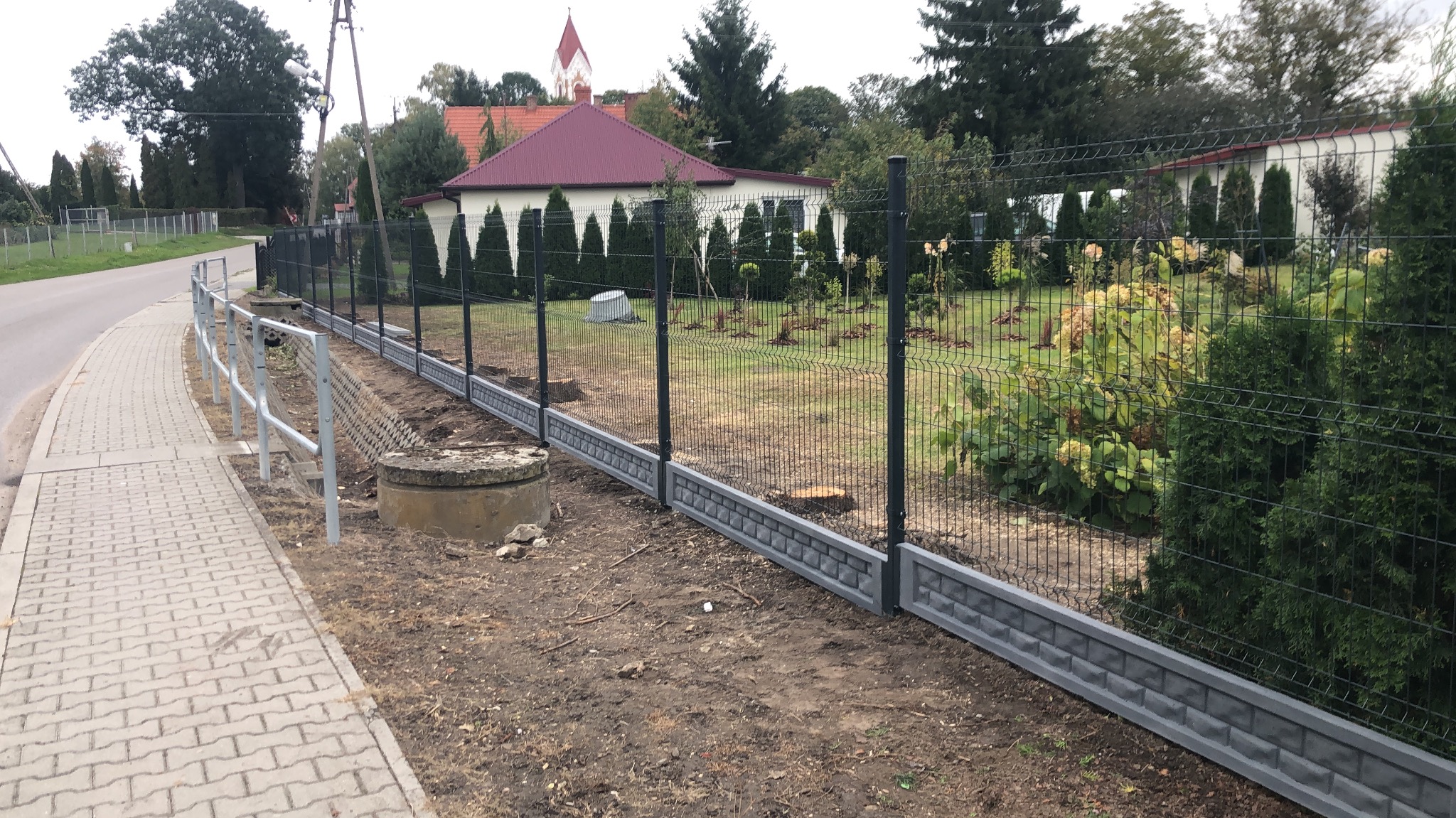 Nowoczesne ogrodzenie panelowe z podmurówką w kolorze grafitowym, oddzielające posesję od chodnika i drogi. W tle widoczny dom z czerwonym dachem i zielenią.