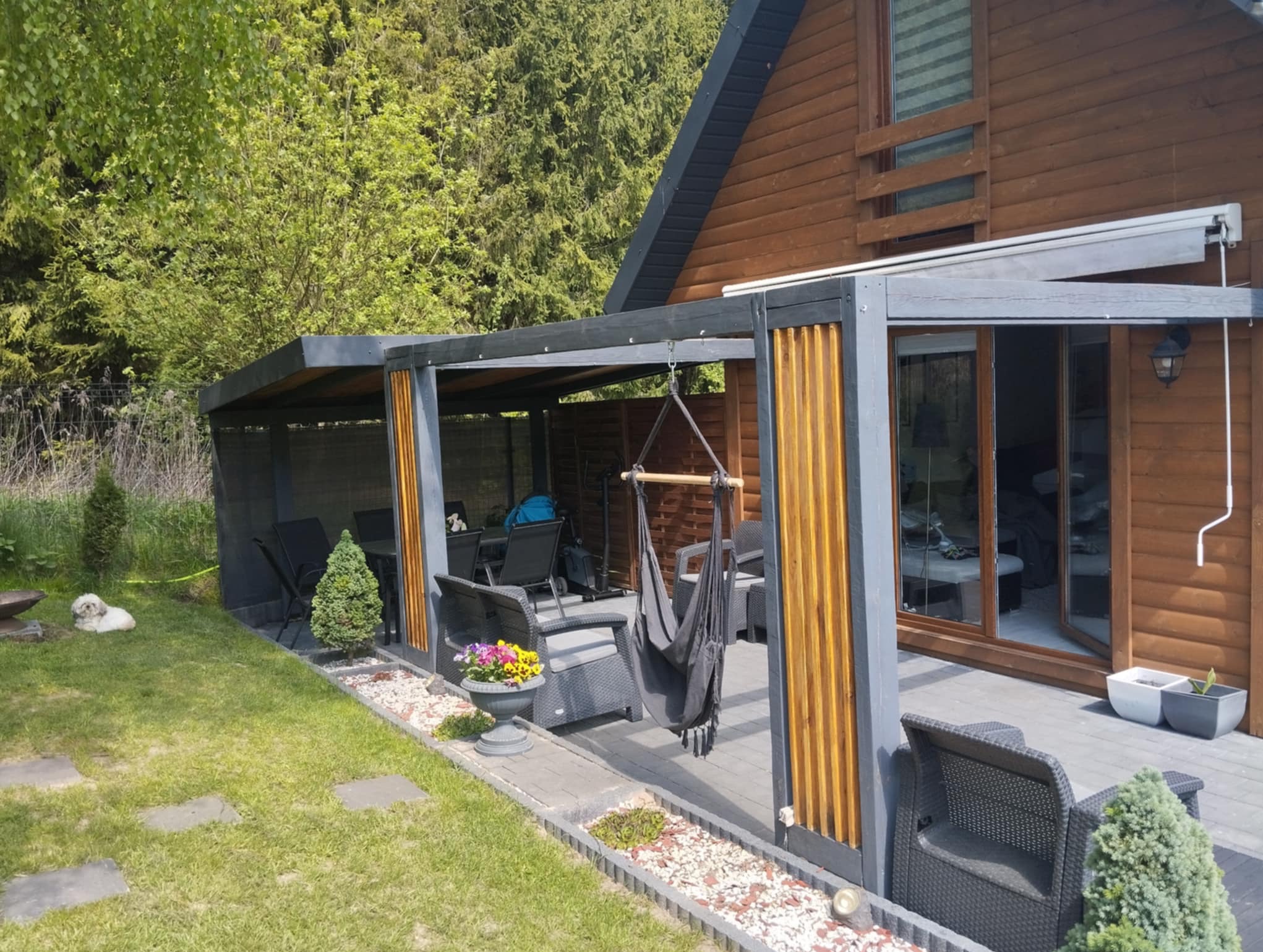 Nowoczesna pergola z szarymi słupami i drewnianymi panelami, wyposażona w wiszący hamak i meble ogrodowe, przylegająca do drewnianego domu z zielonym trawnikiem.