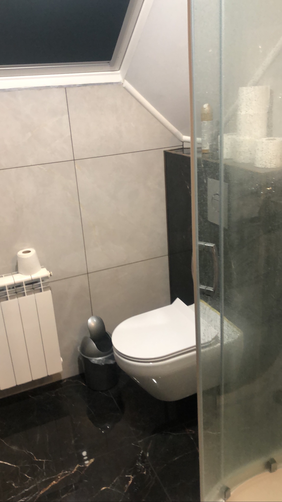 Nowoczesna łazienka z czarną marmurową podłogą, wiszącą toaletą i grzejnikiem. Widoczna kabina prysznicowa z elementami armatury. Uwagę zwraca skośny sufit z oknem.