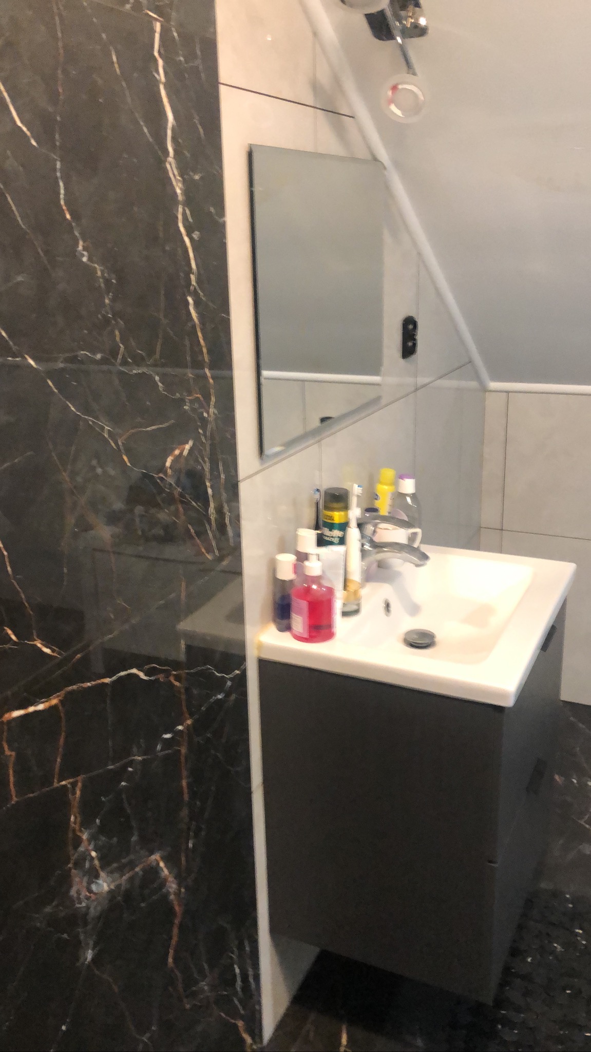 Umywalka z szafką w łazience wykończonej czarnym marmurem z złotymi żyłami. Nad umywalką lustro i oświetlenie punktowe. Widoczny fragment skośnego sufitu.