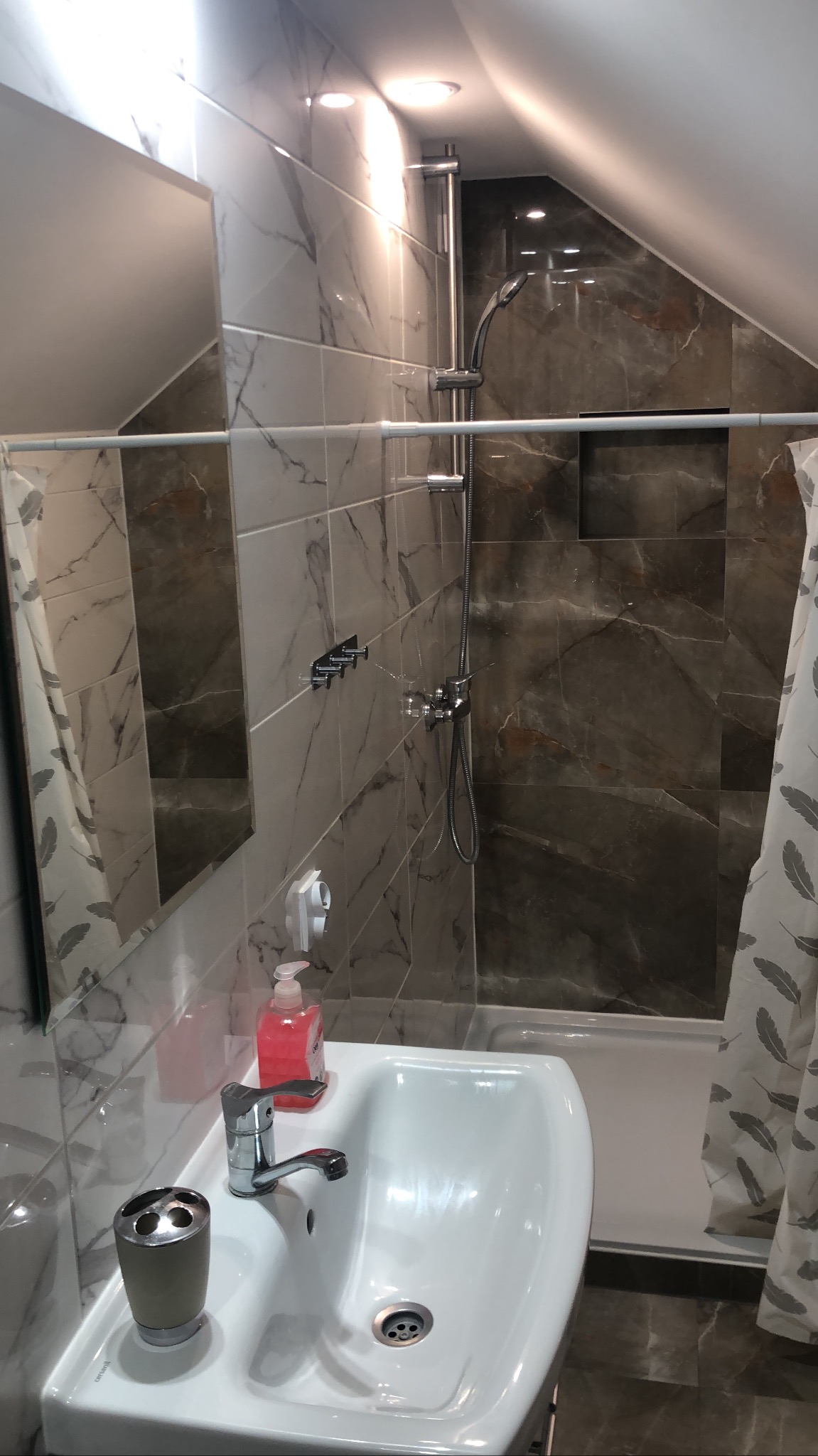 Wykończona łazienka na poddaszu z marmurowymi płytkami, białą umywalką i kabiną prysznicową. Widoczne detale: bateria, dozownik mydła i zasłona prysznicowa.