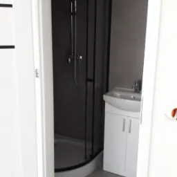 Widok na łazienkę z otwartymi białymi drzwiami, kabina prysznicowa z czarną ramą, biała szafka pod umywalką, szare ściany i podłoga z widocznymi śladami po remoncie.