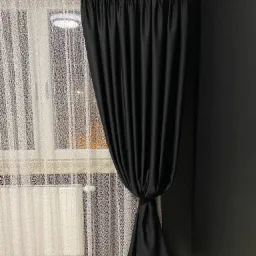 Czarne zasłony zaciemniające, upięte dekoracyjnym węzłem, wiszą obok firanki z delikatnym wzorem na tle okna i ciemnej ściany.