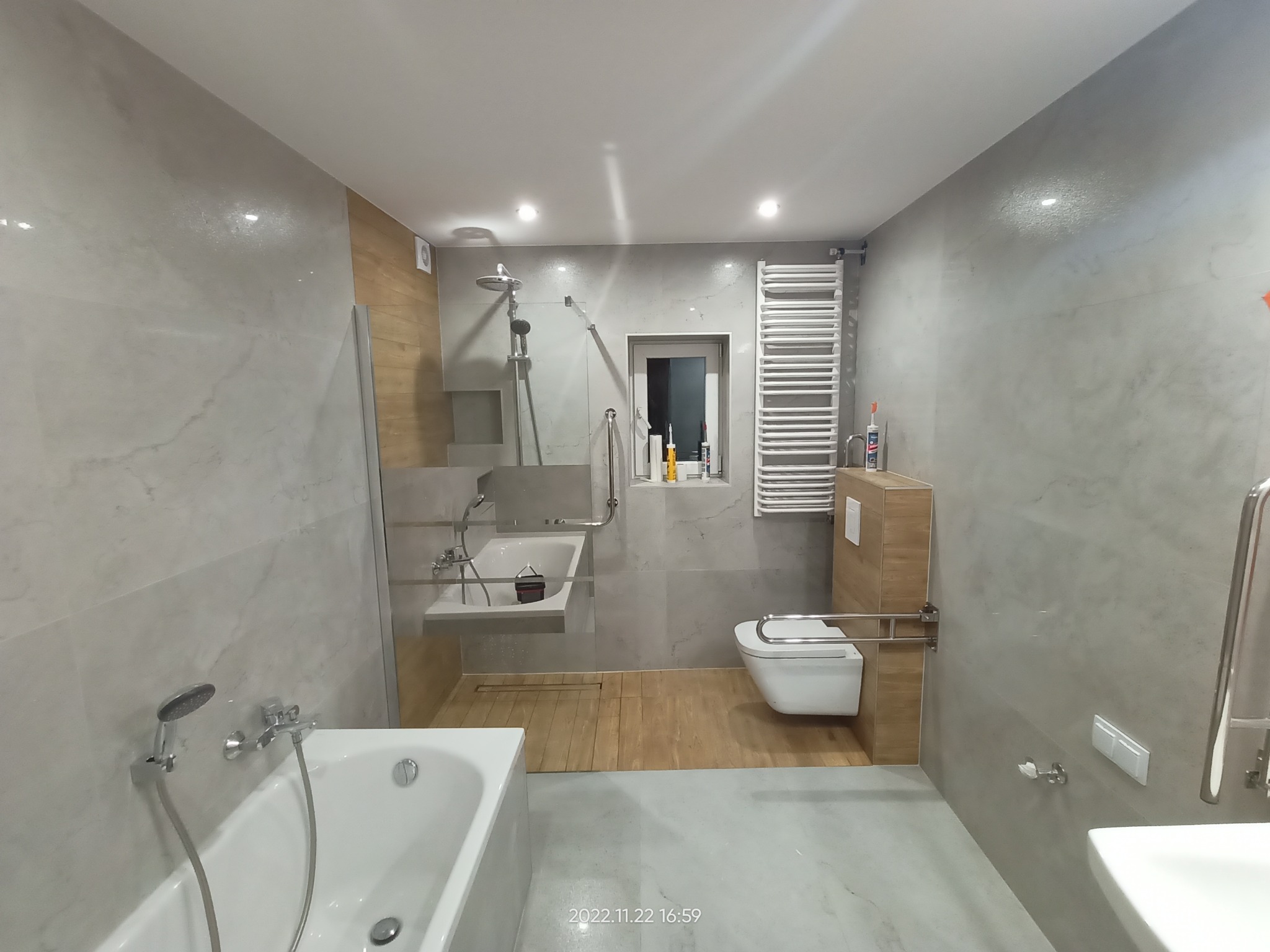 Nowoczesna łazienka z szarymi marmurkowymi płytkami, drewnianymi akcentami i białymi sanitariatami, wyposażona w wannę, umywalkę z półką, toaletę i grzejnik drabinkowy.