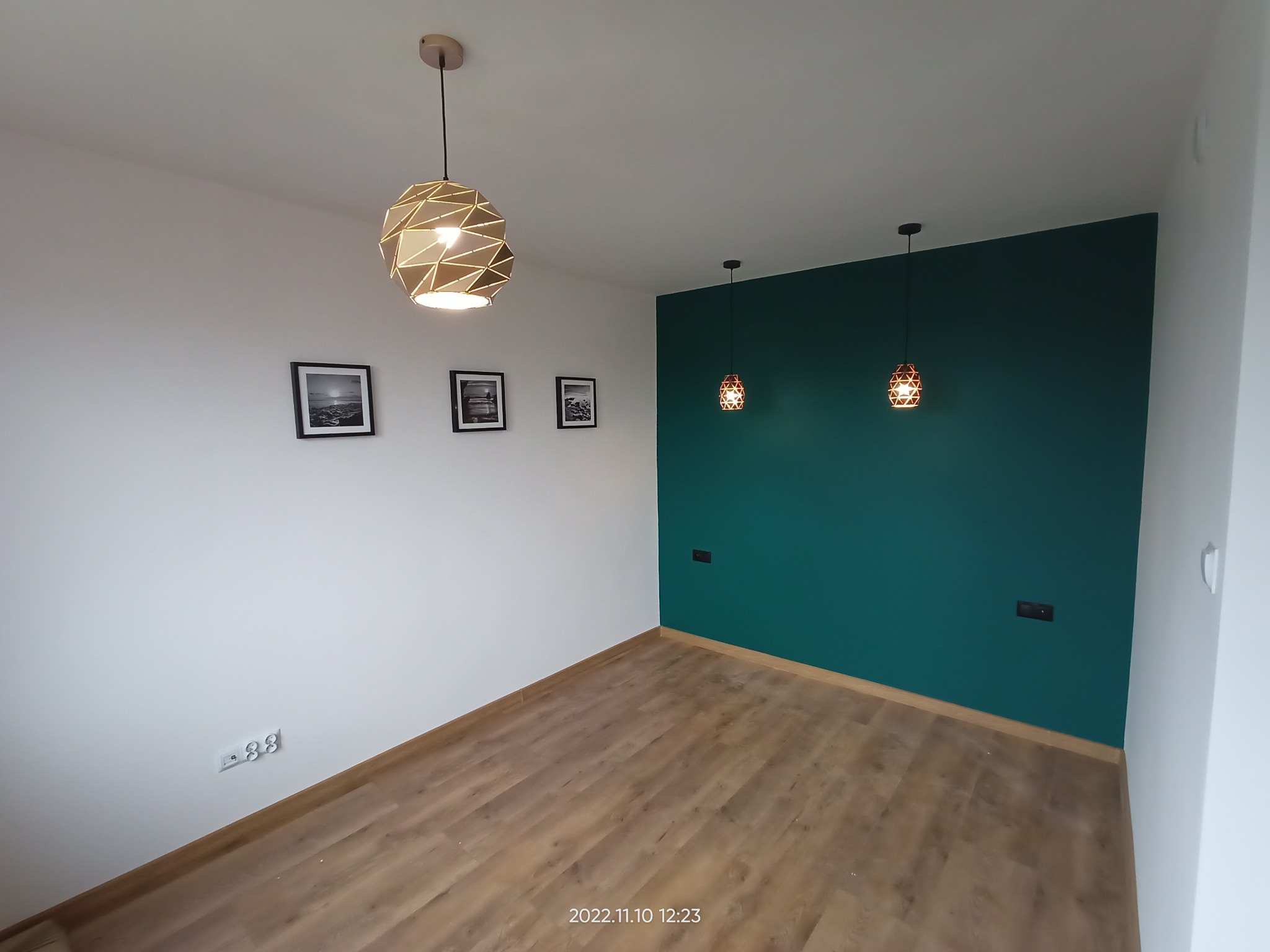 Pomalowane na biało ściany z trzema czarno-białymi obrazkami, drewniane panele podłogowe, ściana akcentująca w odcieniu butelkowej zieleni z dwoma małymi lampami wiszącymi i geometryczną lampą...