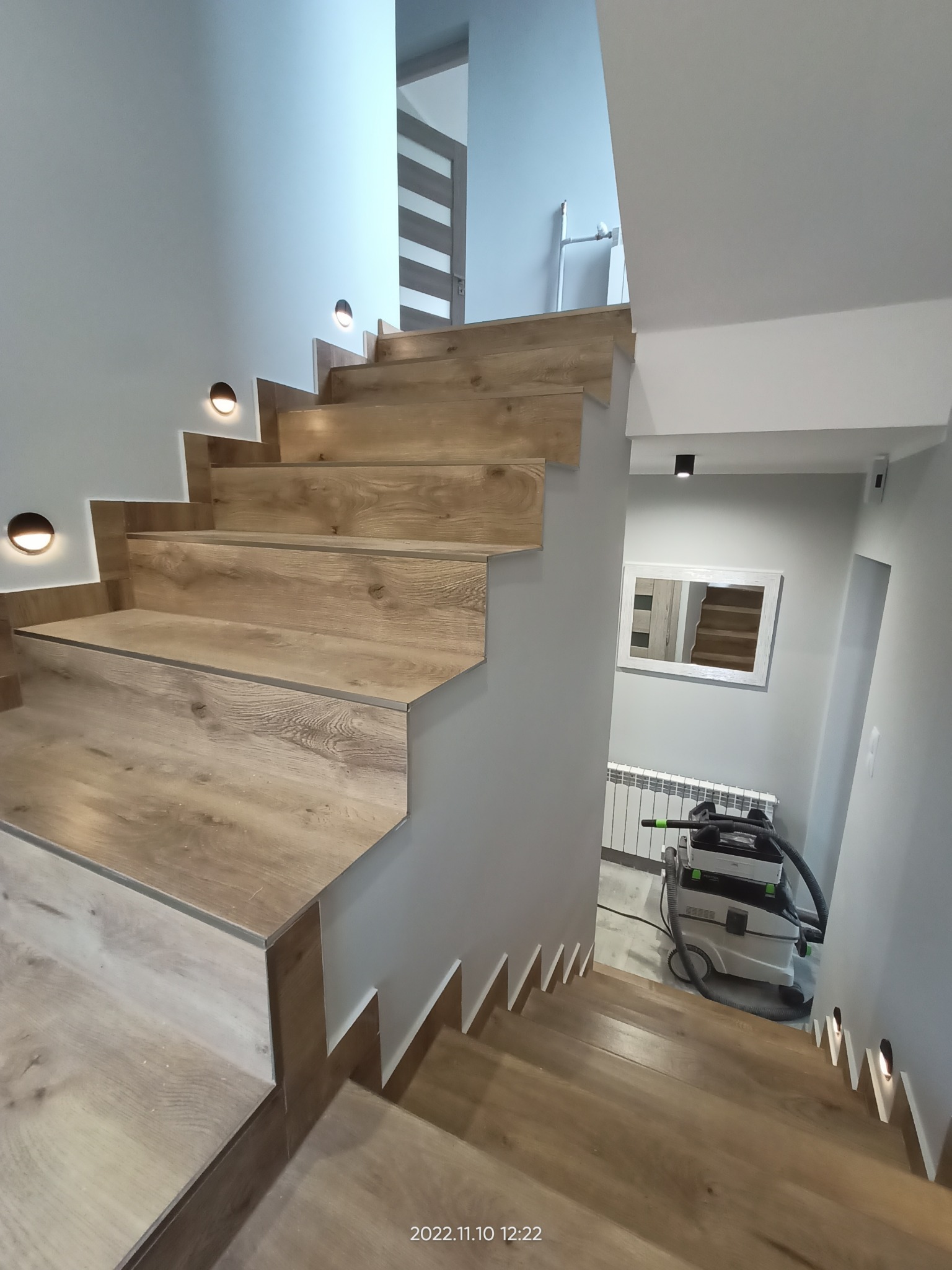 Schody wykończone panelami drewnopodobnymi z oświetleniem punktowym na ścianie, widoczny odkurzacz przemysłowy Festool na podeście.