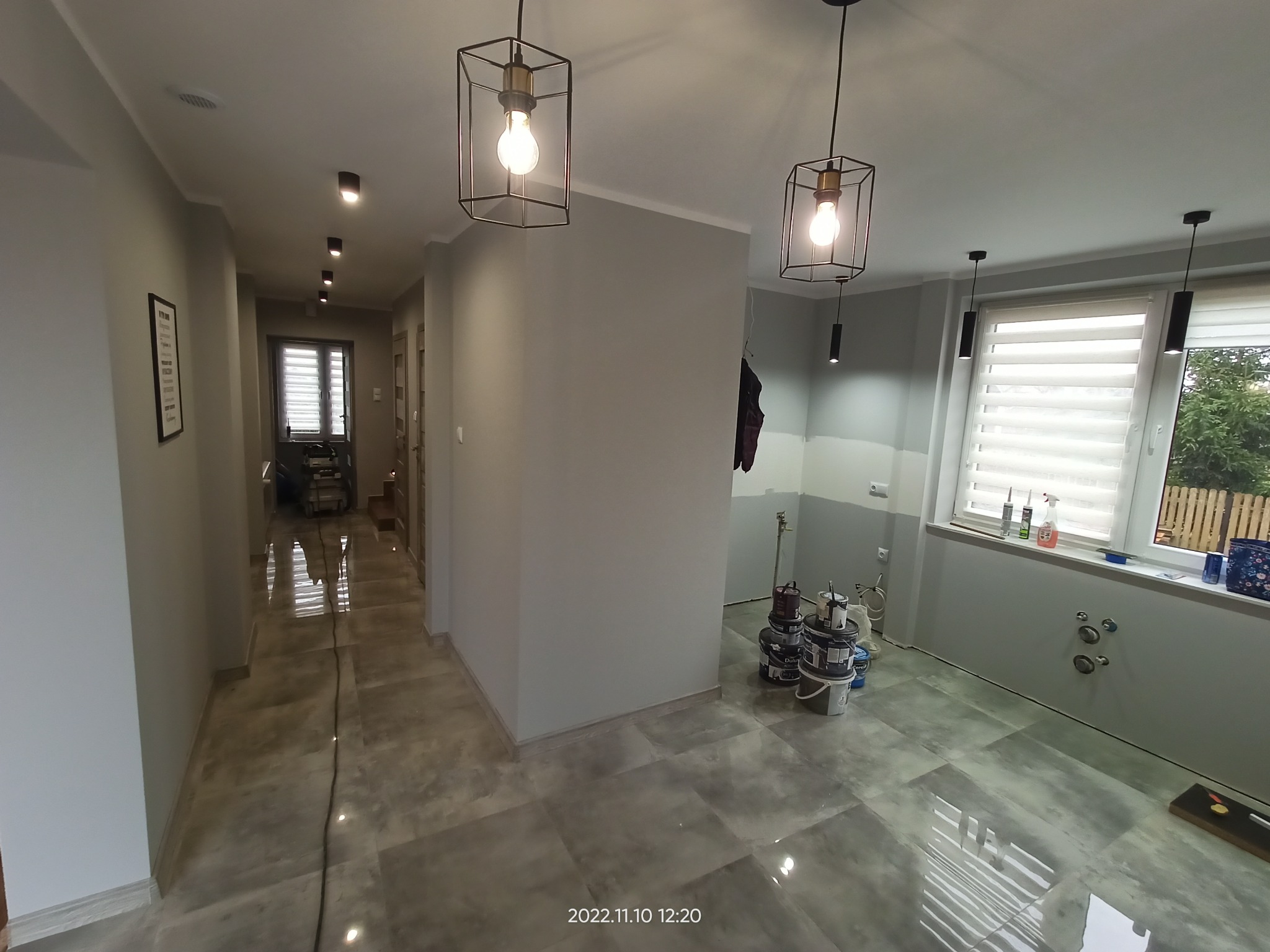 Wnętrze domu w trakcie remontu: szare ściany, błyszcząca podłoga w płytkach, widoczne puszki z farbą i narzędzia malarskie, nowoczesne lampy sufitowe.