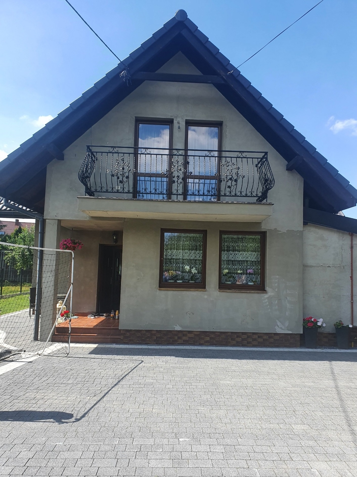 Dom jednorodzinny w trakcie prac elewacyjnych, z widocznym surowym tynkiem, czarną dachówką i ozdobną, kutą balustradą balkonową na pierwszym piętrze. W oknach widoczne firanki i doniczki...