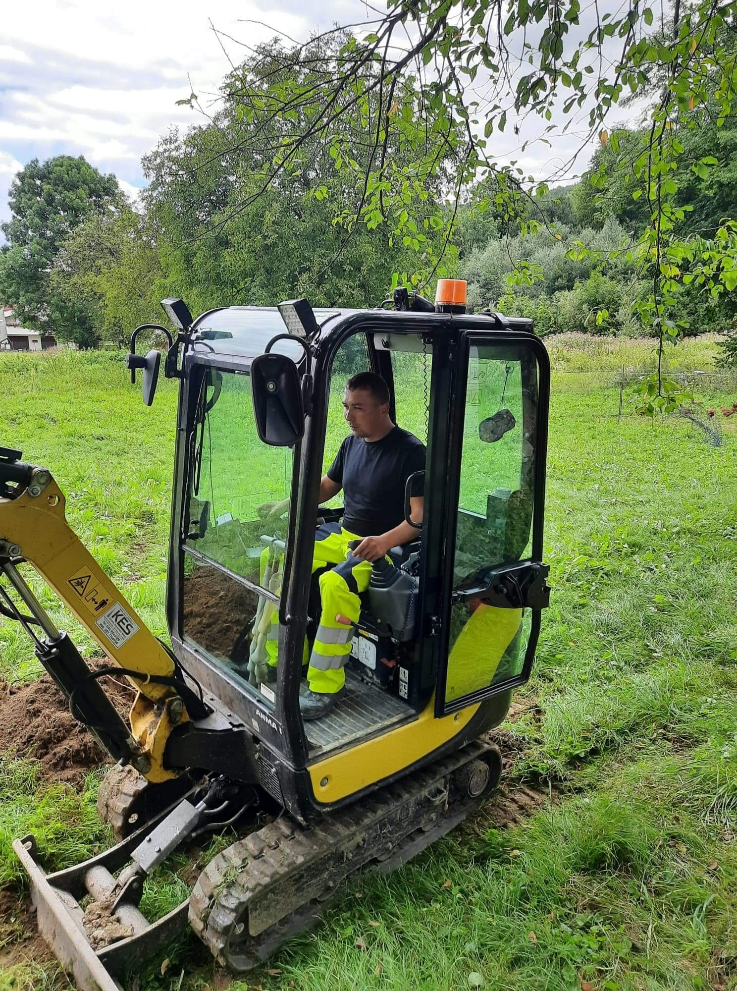 Operator w kabinie żółto-czarnej mini koparki podczas prac ziemnych na zewnątrz, widoczne świeżo wykopane zagłębienie w trawie.