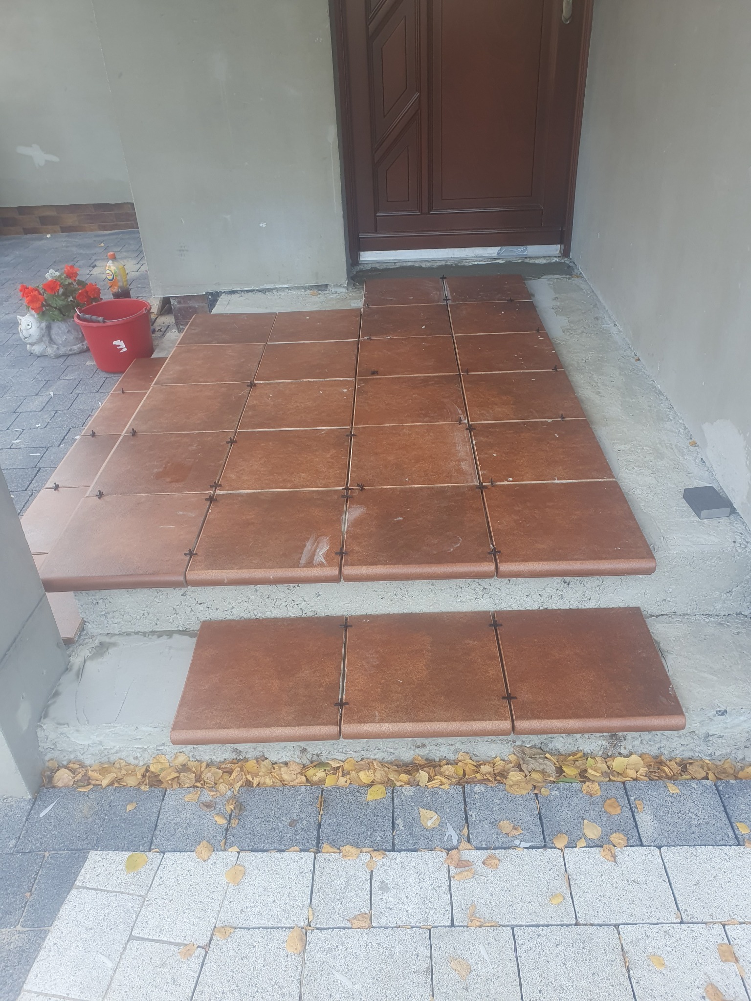 Montaż brązowych płytek ceramicznych na schodach wejściowych, widoczne krzyżyki dystansowe, obok wiadro i kwiaty w doniczce.
