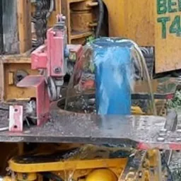 Wiercenie studni: Woda tryska z otworu wiertniczego podczas procesu wiercenia, widoczna część maszyny wiertniczej z żółtymi elementami i łańcuchem. Niebieska rura w strumieniu wody.