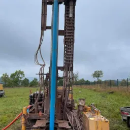 Wiertnica do studni głębinowej w Krosnowicach, widok na maszynę w trakcie pracy z wystającą niebieską rurą, błotniste podłoże i zalesione wzgórza w tle.