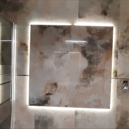 Grzegorz Śledź - Wykończona łazienka z kabiną prysznicową typu walk-in, podświetlanymi płytkami imitującymi marmur i widocznym fragmentem niezakończonego wykończenia ściany z surowym betonem.