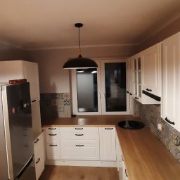 Grzegorz Śledź - Wykończona kuchnia z białymi szafkami, drewnianym blatem, panelami podłogowymi i czarną lampą wiszącą, widok z perspektywy szerokokątnej.