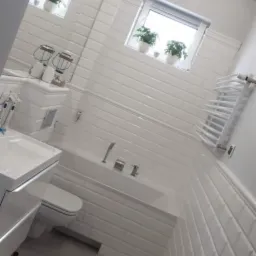 Biała łazienka po remoncie, z wanną obłożoną kafelkami typu metro, umywalką z szafką, toaletą, grzejnikiem drabinkowym i dwoma oknami z roślinami w doniczkach.