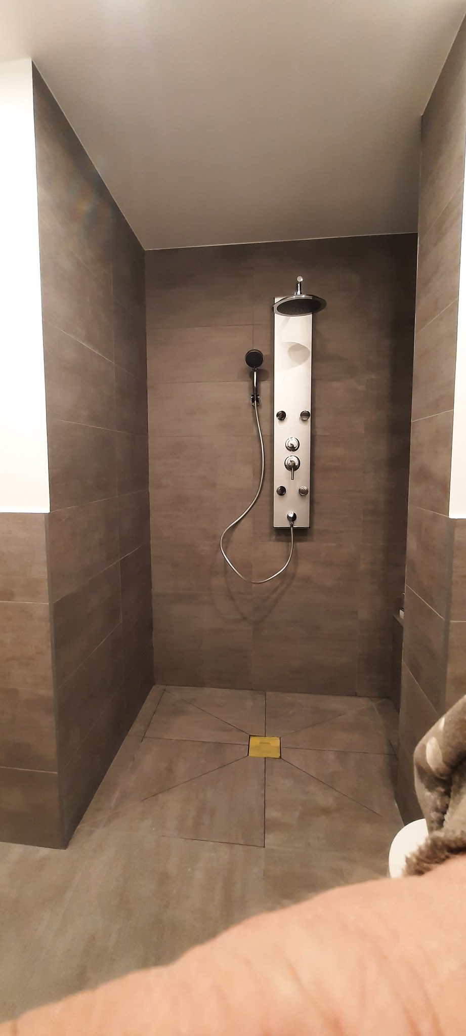 Nowoczesna kabina prysznicowa z panelem natryskowym, wykończona płytkami w odcieniach szarości, z centralnie umieszczonym odpływem podłogowym w kolorze złotym.