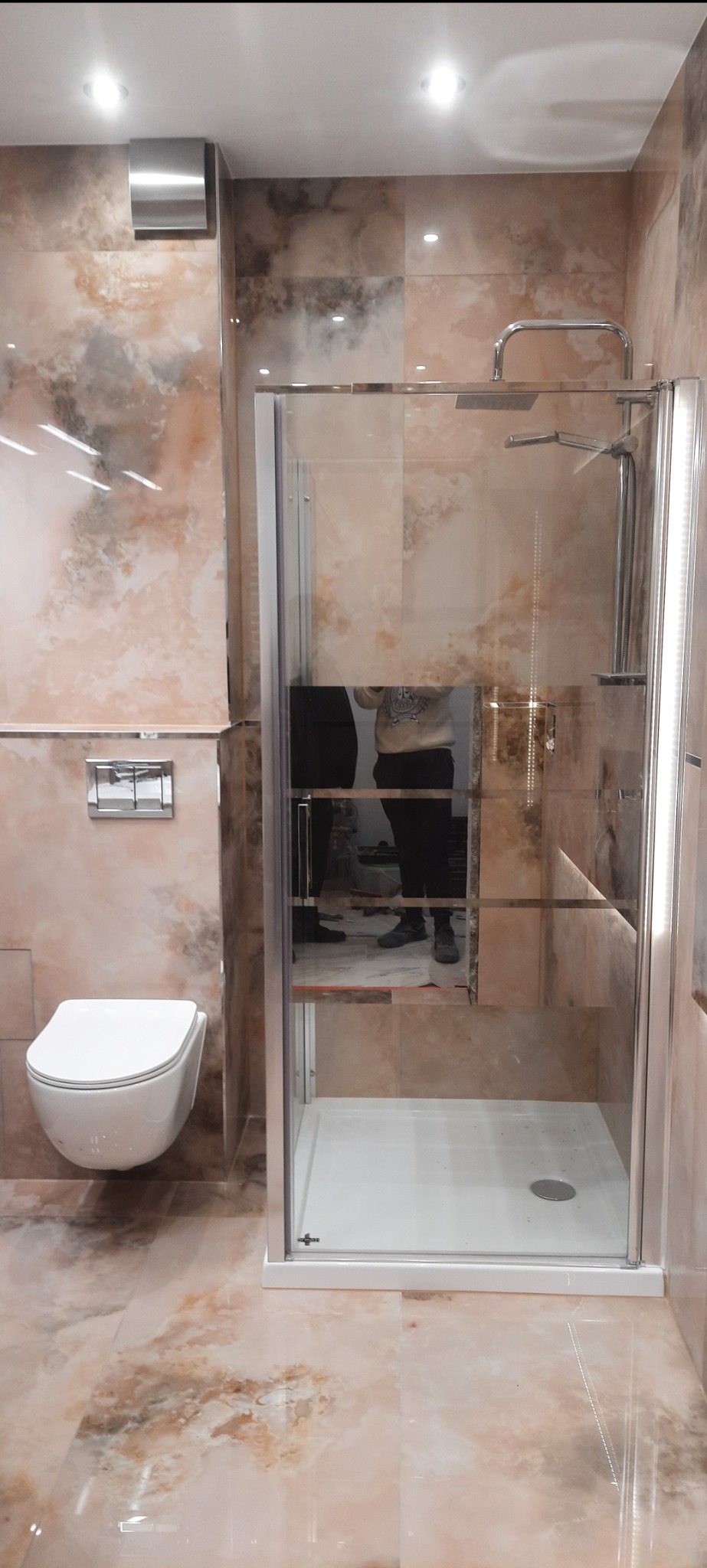 Nowoczesna kabina prysznicowa z chromowanymi elementami, zamontowana w łazience wykończonej płytkami imitującymi marmur, z podwieszaną toaletą i widocznym odbiciem osoby w szkle.