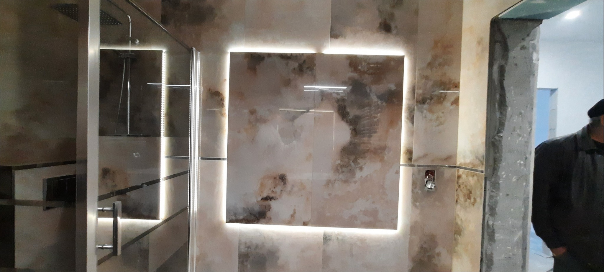 Wykończona łazienka z kabiną prysznicową typu walk-in, podświetlanymi płytkami imitującymi marmur i widocznym fragmentem niezakończonego wykończenia ściany z surowym betonem.