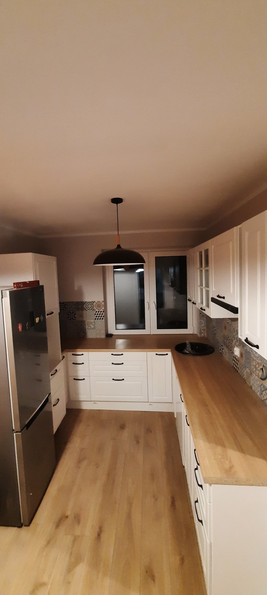 Wykończona kuchnia z białymi szafkami, drewnianym blatem, panelami podłogowymi i czarną lampą wiszącą, widok z perspektywy szerokokątnej.