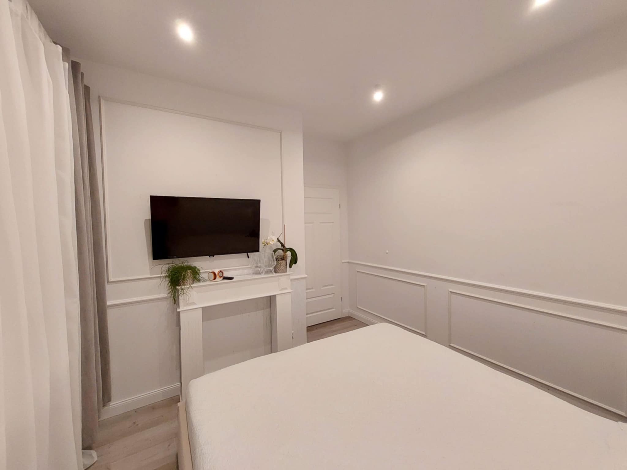 Jasna sypialnia z białymi ścianami zdobionymi sztukaterią, białe zasłony, telewizor na ścianie nad imitacją kominka z roślinami i dekoracjami, oraz duże łóżko z białą pościelą.