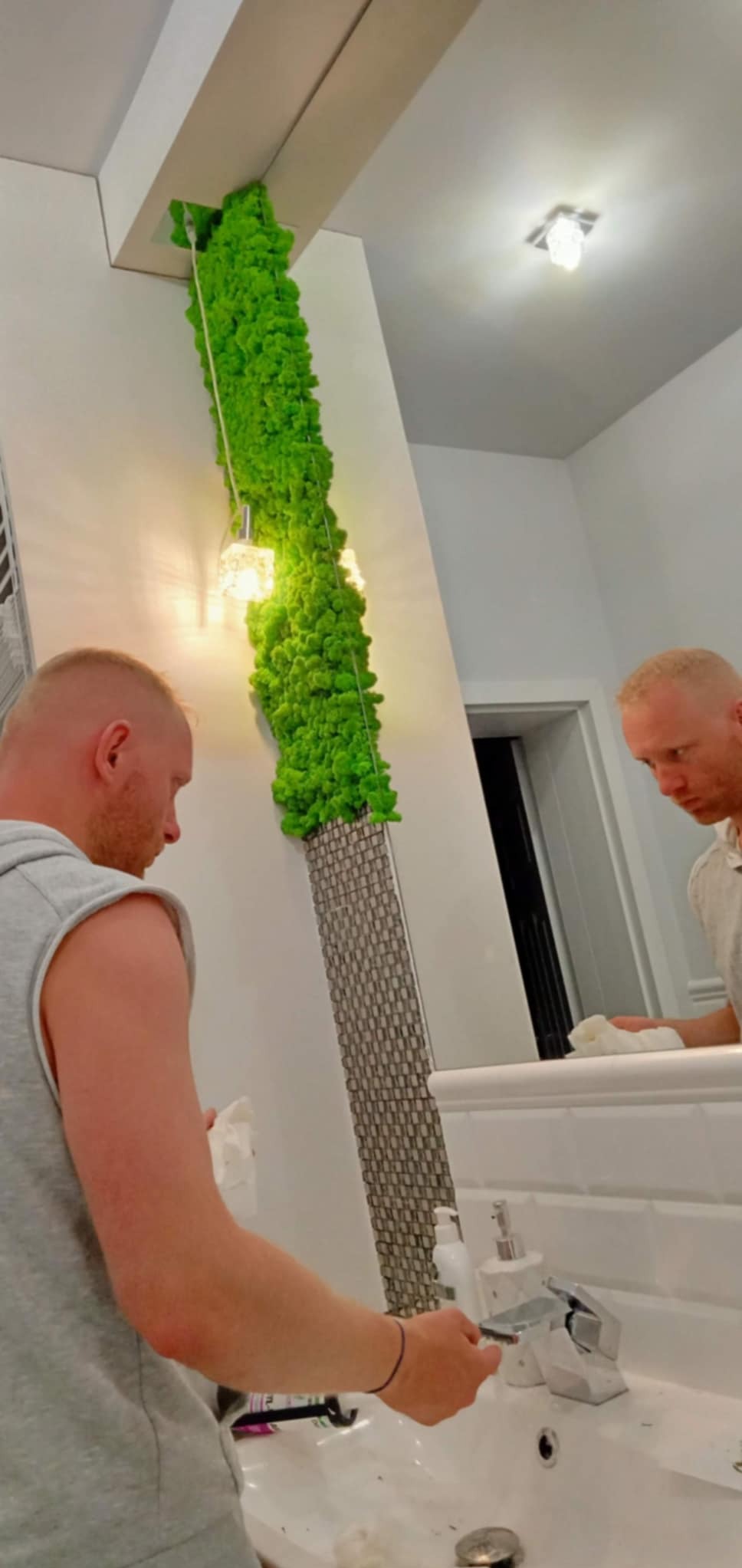 Wnętrze łazienki z dekoracyjną ścianą z mchu, oświetloną dwoma lampkami, odbijającą się w lustrze nad umywalką, gdzie osoba czyści powierzchnię.