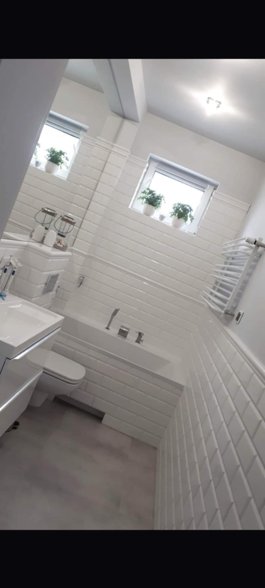 Biała łazienka po remoncie, z wanną obłożoną kafelkami typu metro, umywalką z szafką, toaletą, grzejnikiem drabinkowym i dwoma oknami z roślinami w doniczkach.