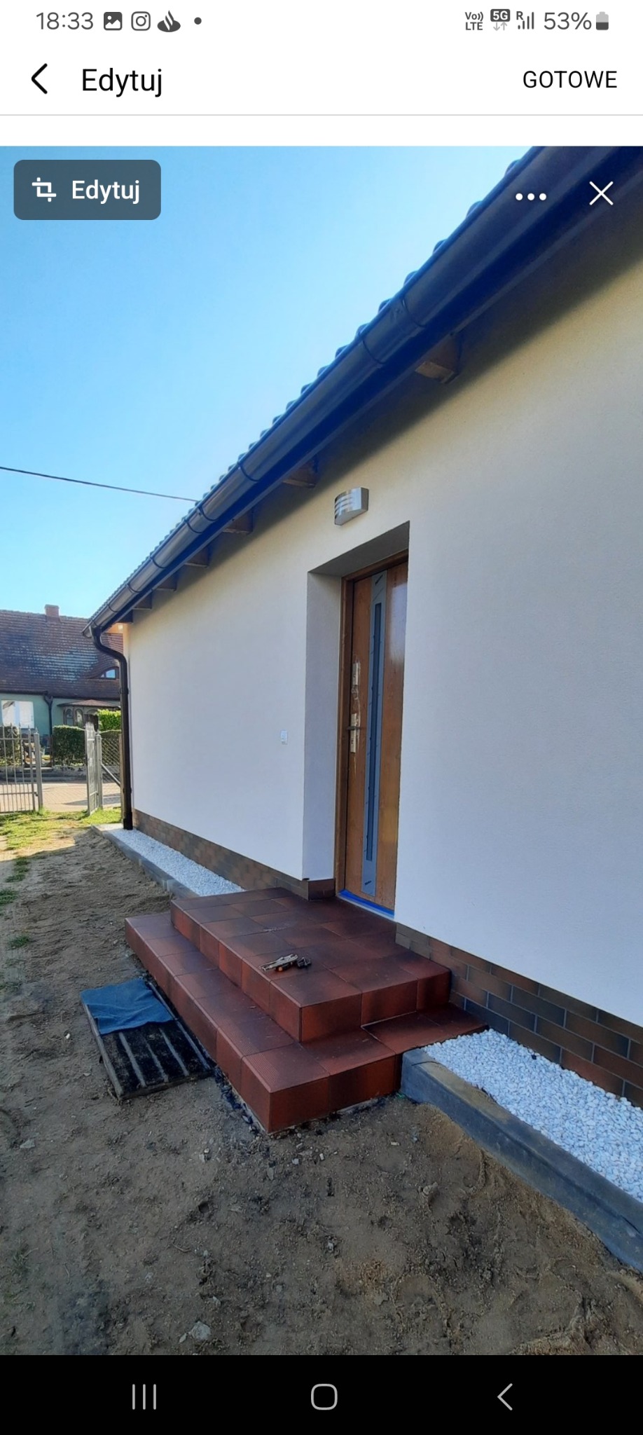 Nowe, brązowe płytki schodowe prowadzące do drzwi wejściowych domu z jasną elewacją, widoczne narzędzie na schodach i ozdobny pas z białego żwiru przy ścianie budynku.