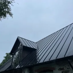 Czarny, metalowy dach z oknem dachowym na kamiennym budynku z klimatyzatorem na ścianie, widok z dołu w pochmurny dzień.