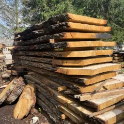 Stos desek z surowego drewna ułożonych na zewnątrz, z widoczną korą i naturalnymi krawędziami, przygotowanych do obróbki lub wykorzystania w budowie domu szkieletowego.