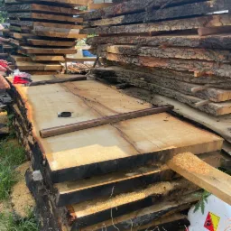 Stos desek z surowego drewna z korą, przygotowanych do obróbki, leżących na trawie obok pojemnika z symbolem ostrzegawczym.