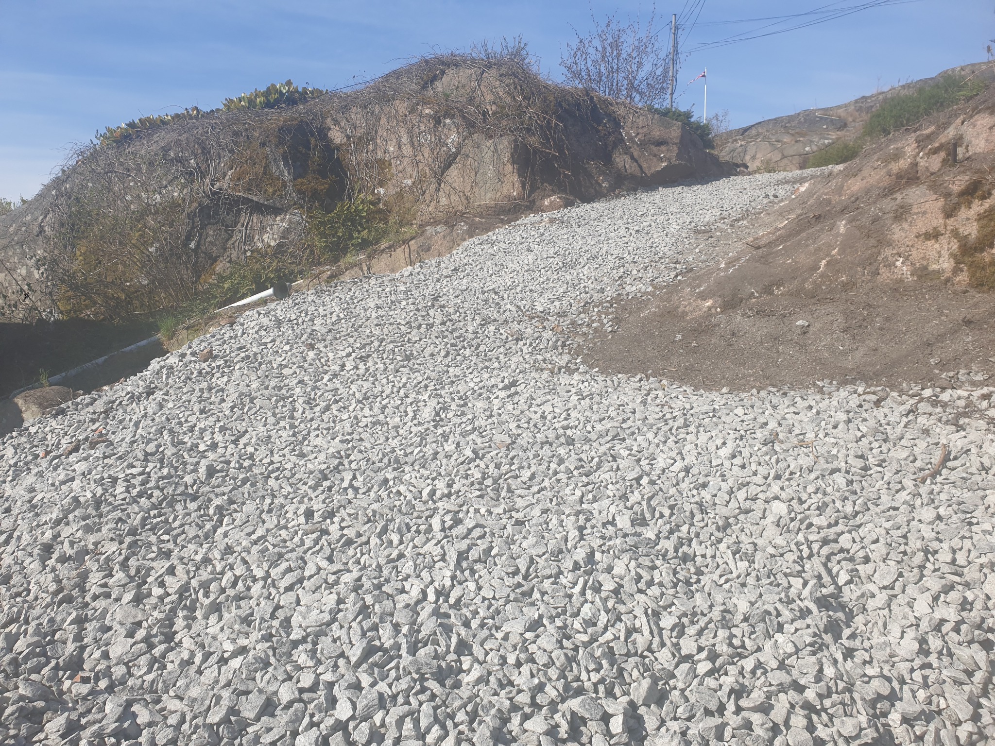 Ukształtowany teren z wysypanym drobnym żwirem, skarpa z kamieniami i roślinnością w tle pod błękitnym niebem.