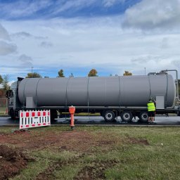 Concret - Szary samochód ciężarowy z cysterną na parkingu, pracownik w żółtej kamizelce otwiera właz. Widoczne zabezpieczenia budowlane i częściowo rozkopana ziemia.