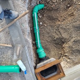 Concret - Zielona rura kanalizacyjna w wykopie, podłączona do studzienki rewizyjnej, widoczne narzędzia i fragment obuwia pracownika, ziemia i kostka brukowa w tle.