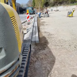 Żółta minikoparka Wacker Neuson na placu budowy, obok ułożone krawężniki z kostki, w tle pracownicy i zagęszczarka wibracyjna.