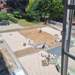 Concret - Widok z góry na plac budowy z częściowo ułożoną kostką brukową w odcieniach beżu i brązu, pracownik wyrównuje piasek pod dalsze układanie, palety z kostką czekają na ułożenie, w tle budynki i zieleń.