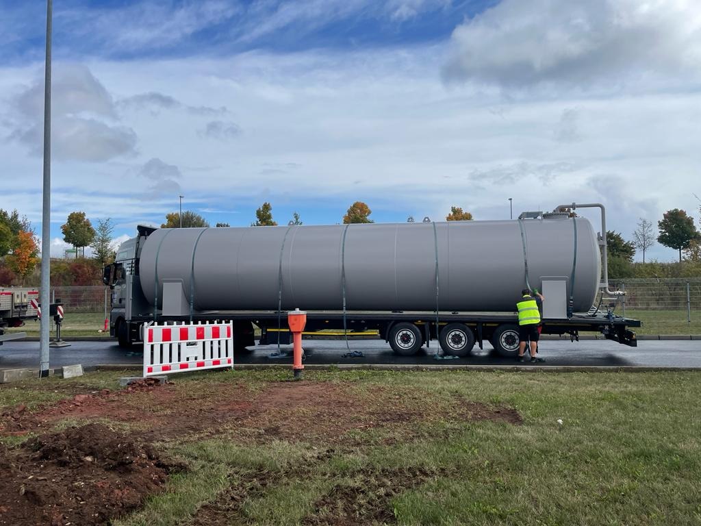 Szary samochód ciężarowy z cysterną na parkingu, pracownik w żółtej kamizelce otwiera właz. Widoczne zabezpieczenia budowlane i częściowo rozkopana ziemia.
