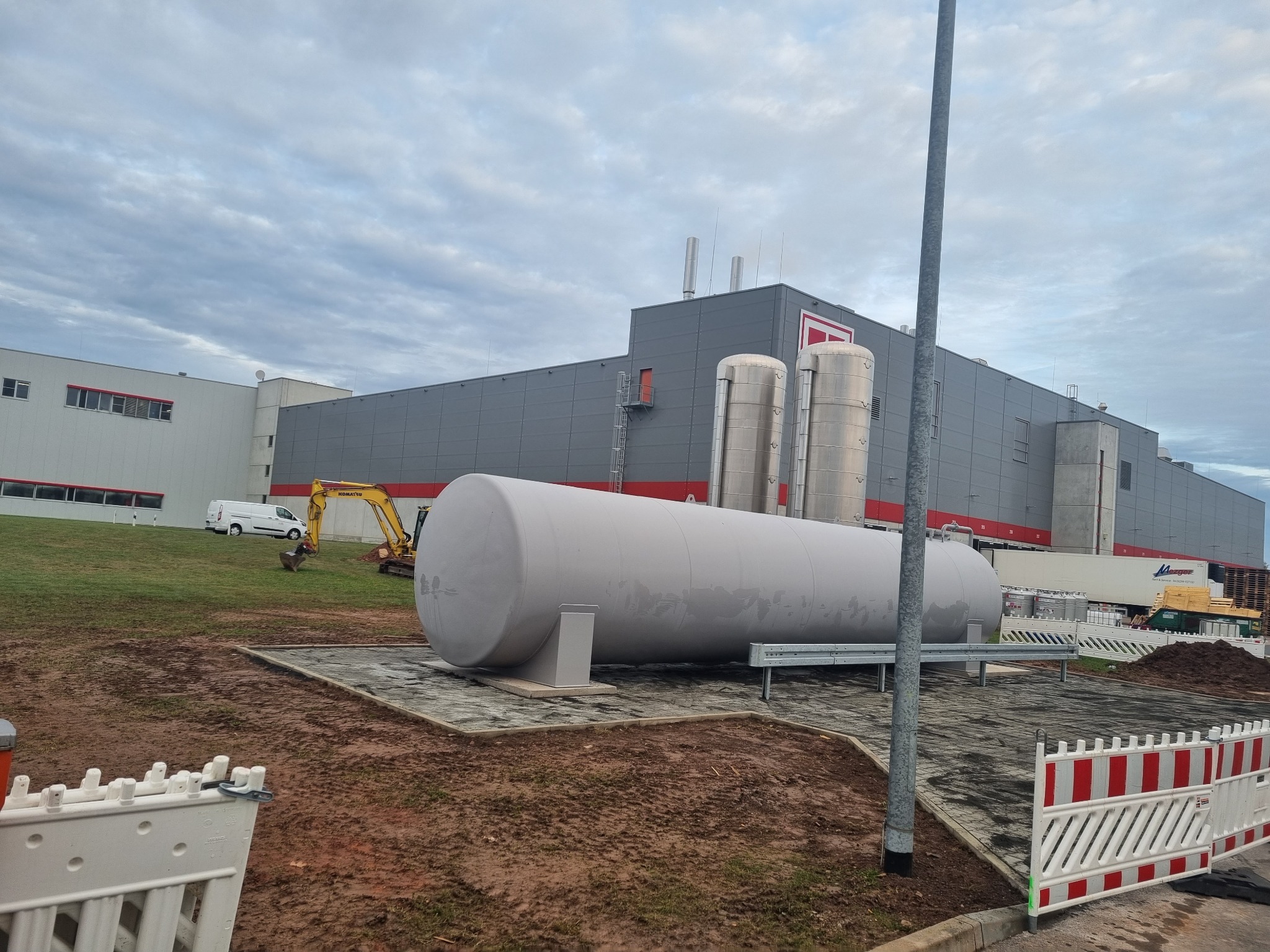 Duży, szary zbiornik cylindryczny na betonowej podstawie, w tle budynek przemysłowy z dwoma srebrnymi silosami i koparka wykonująca wykop, pochmurne niebo.