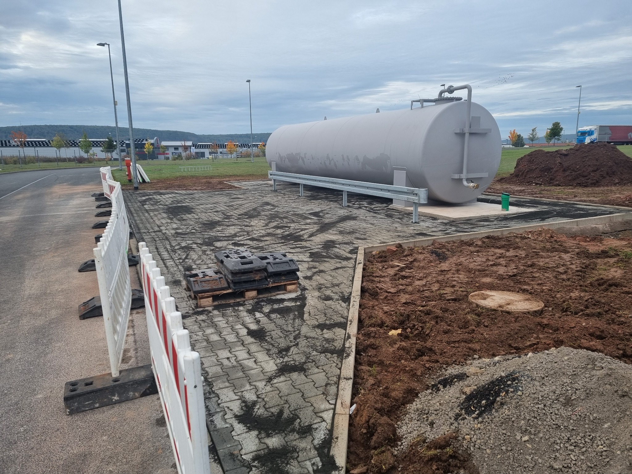 Szary, poziomy zbiornik na podwyższeniu z metalową barierką, umieszczony na placu wyłożonym kostką brukową, w tle widoczna ziemia, ogrodzenie i budynki.