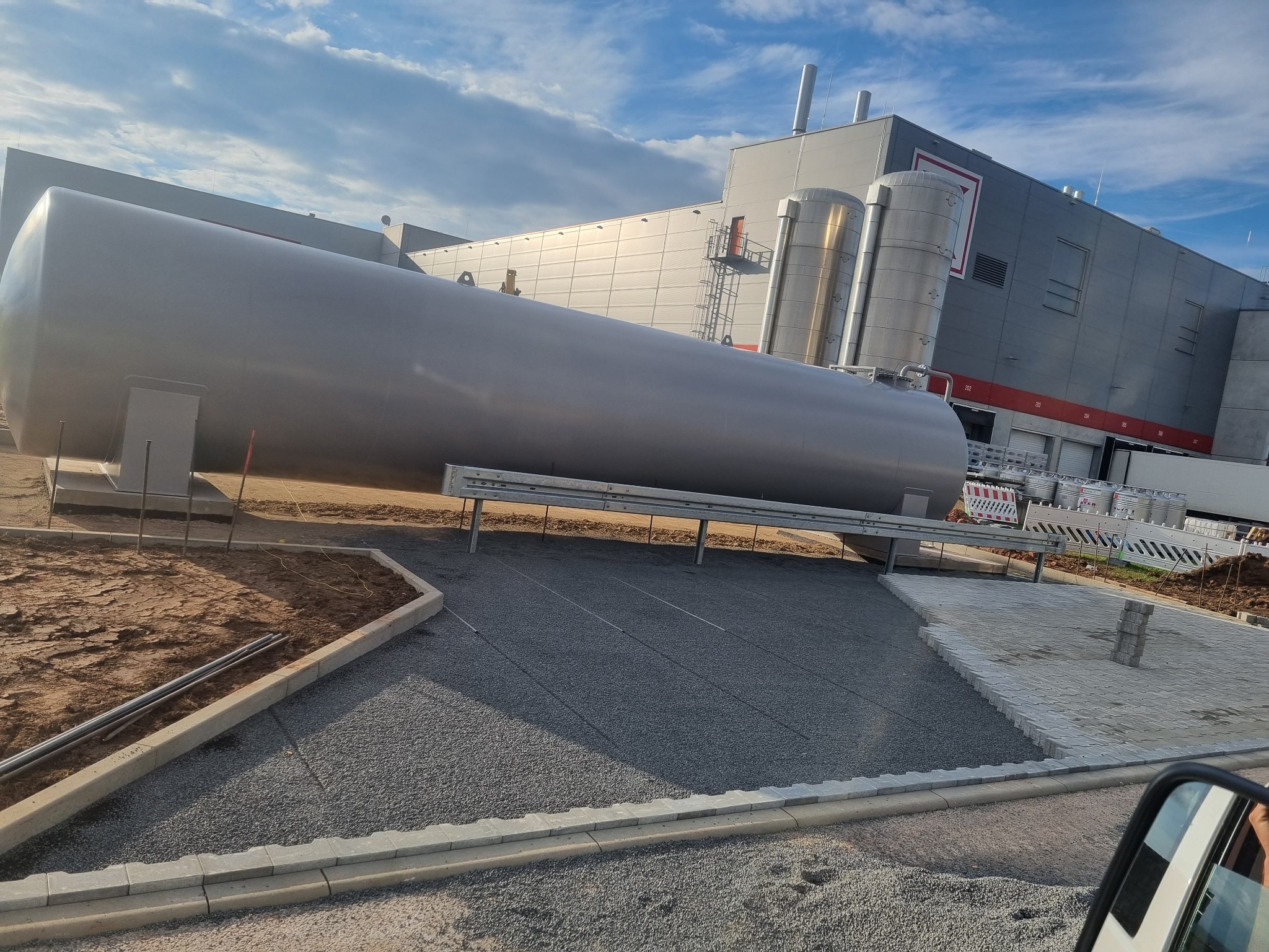 Poziomy, srebrny zbiornik na fundamencie z kruszywa obok budynku przemysłowego z metalowymi kominami i rampą załadunkową, widoczne przygotowania do wykończenia terenu.