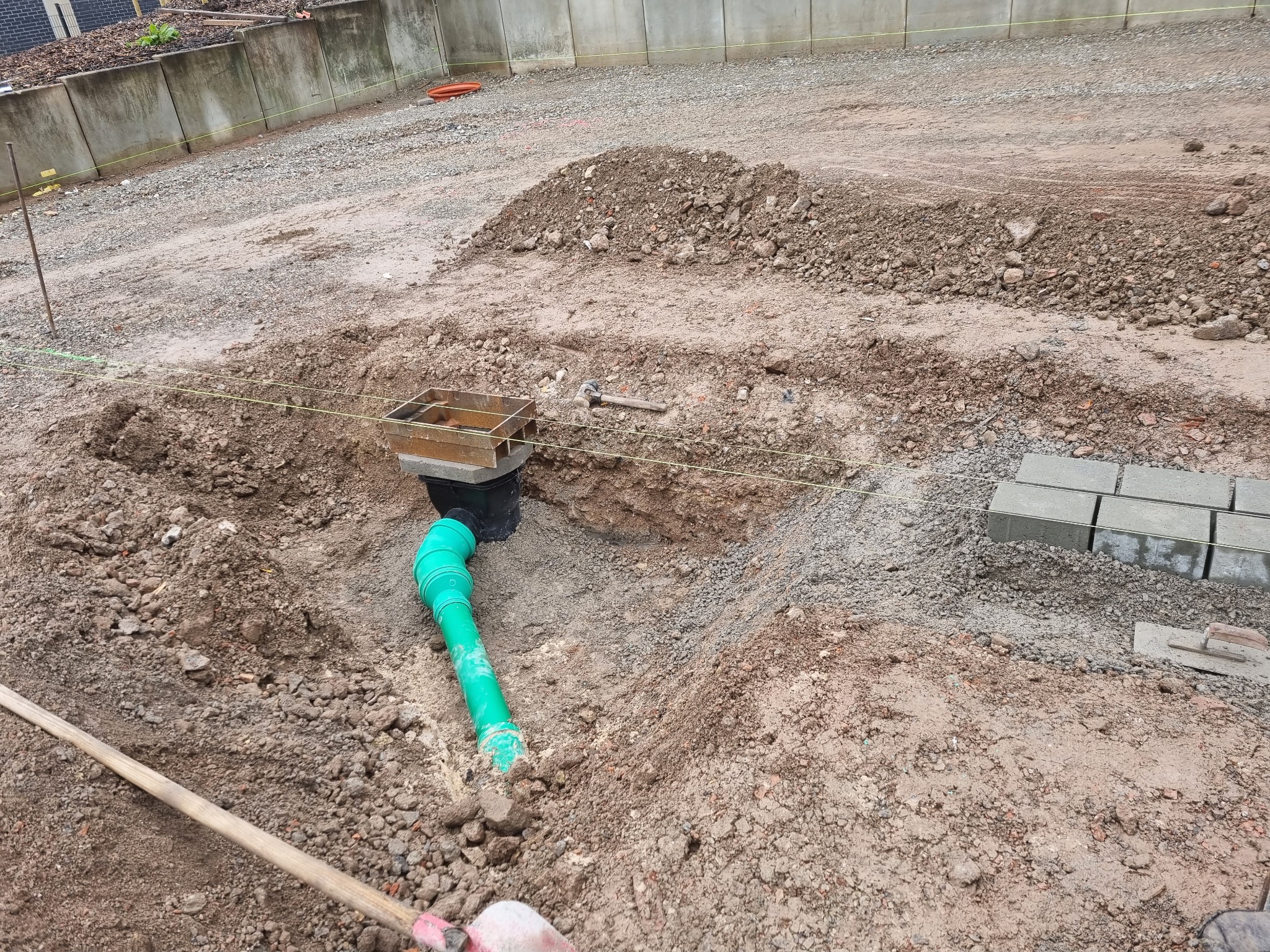 Wykop z zieloną rurą kanalizacyjną i metalową skrzynką, przygotowany pod przyłącze, z widocznym sznurkiem wyznaczającym linię, narzędziami i kostką brukową w tle.