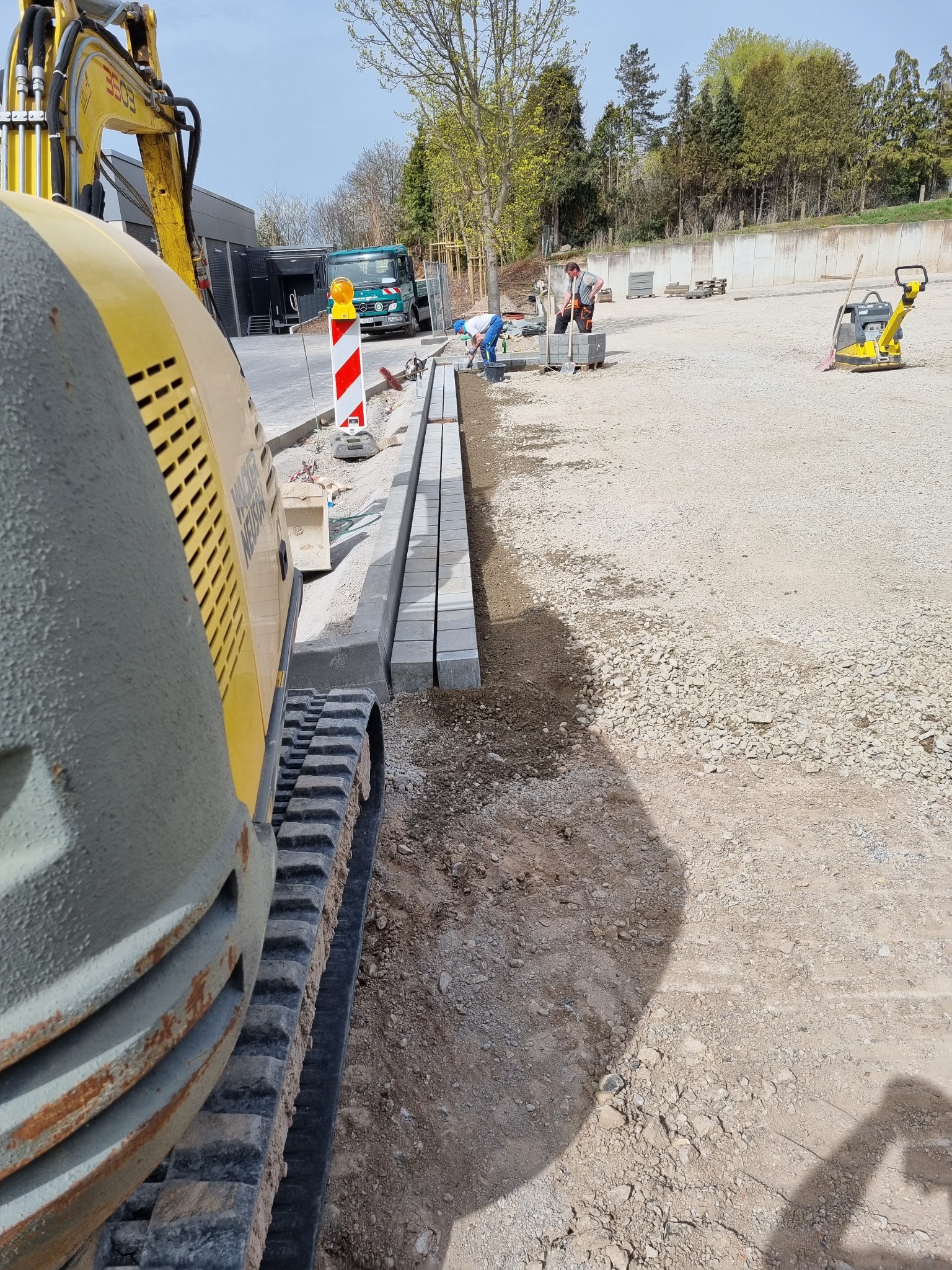 Żółta minikoparka Wacker Neuson na placu budowy, obok ułożone krawężniki z kostki, w tle pracownicy i zagęszczarka wibracyjna.