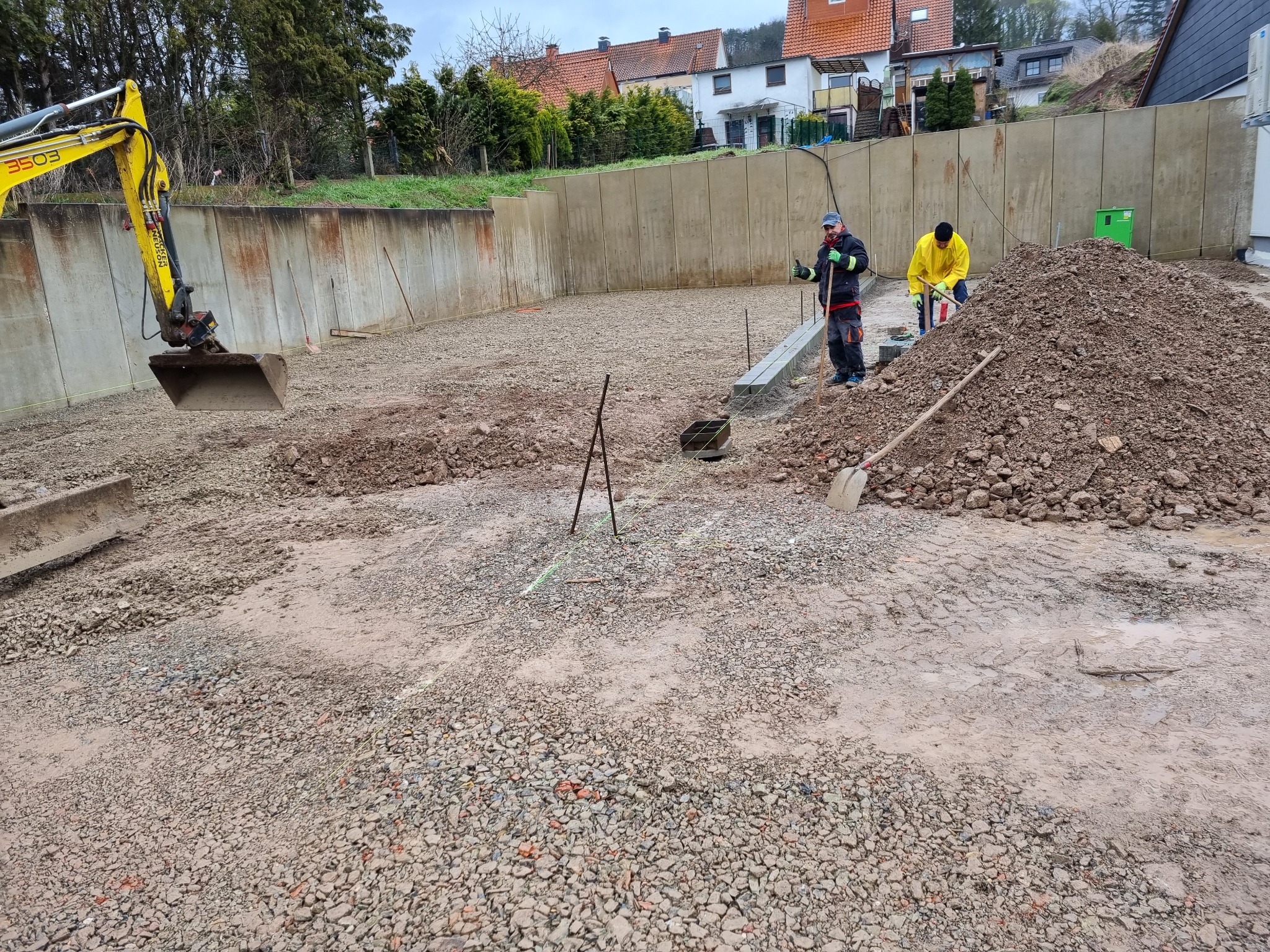 Przygotowanie terenu pod budowę: robotnicy z łopatami, mini koparka i wytyczone fundamenty na żwirowym podłożu, w tle betonowy mur oporowy i zabudowa jednorodzinna.