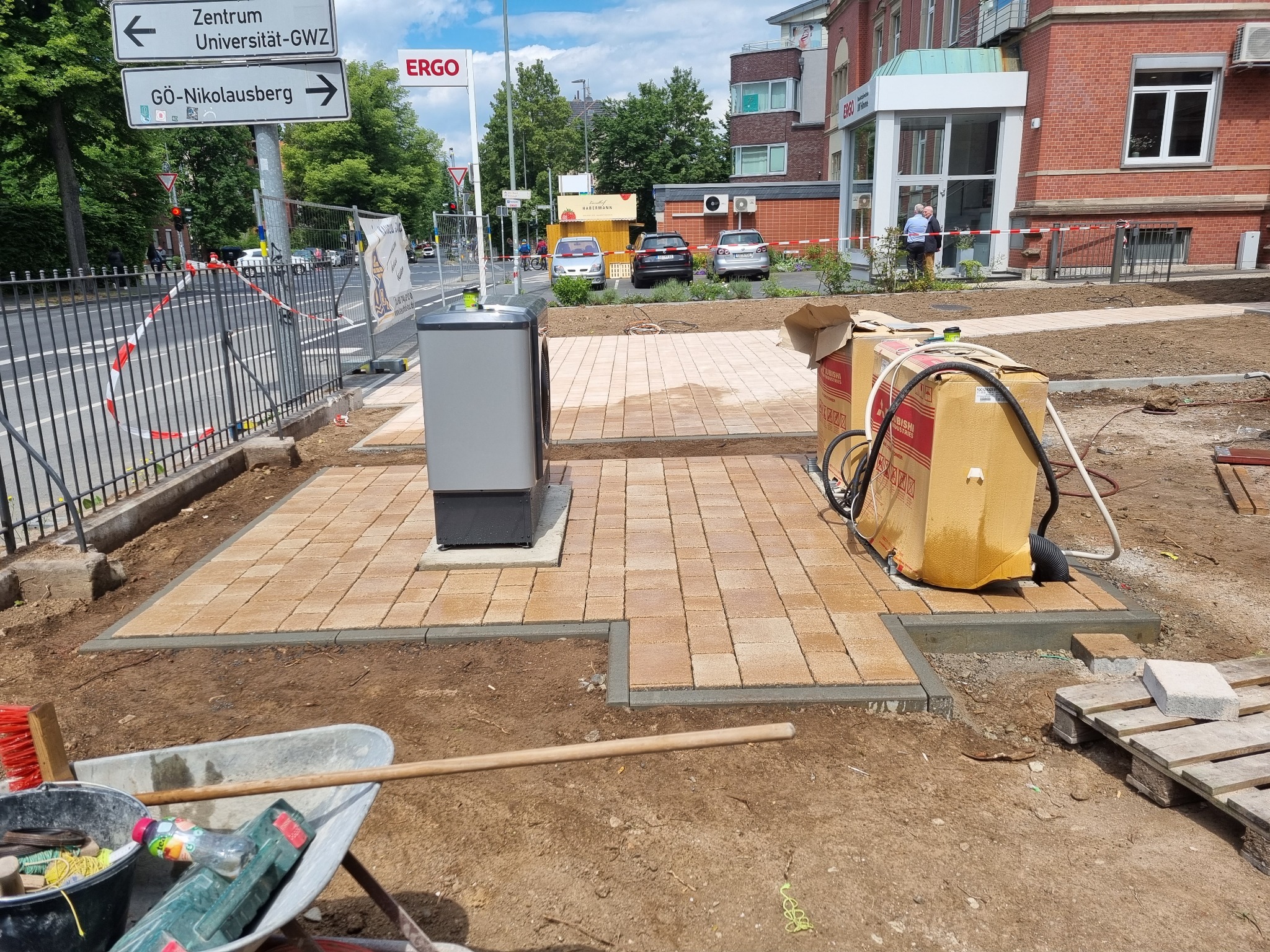 Fragment nowej nawierzchni z kostki brukowej w odcieniach beżu, otoczona ziemią i narzędziami budowlanymi, z widocznym urządzeniem technicznym po lewej stronie oraz kartonami po prawej, w tle...