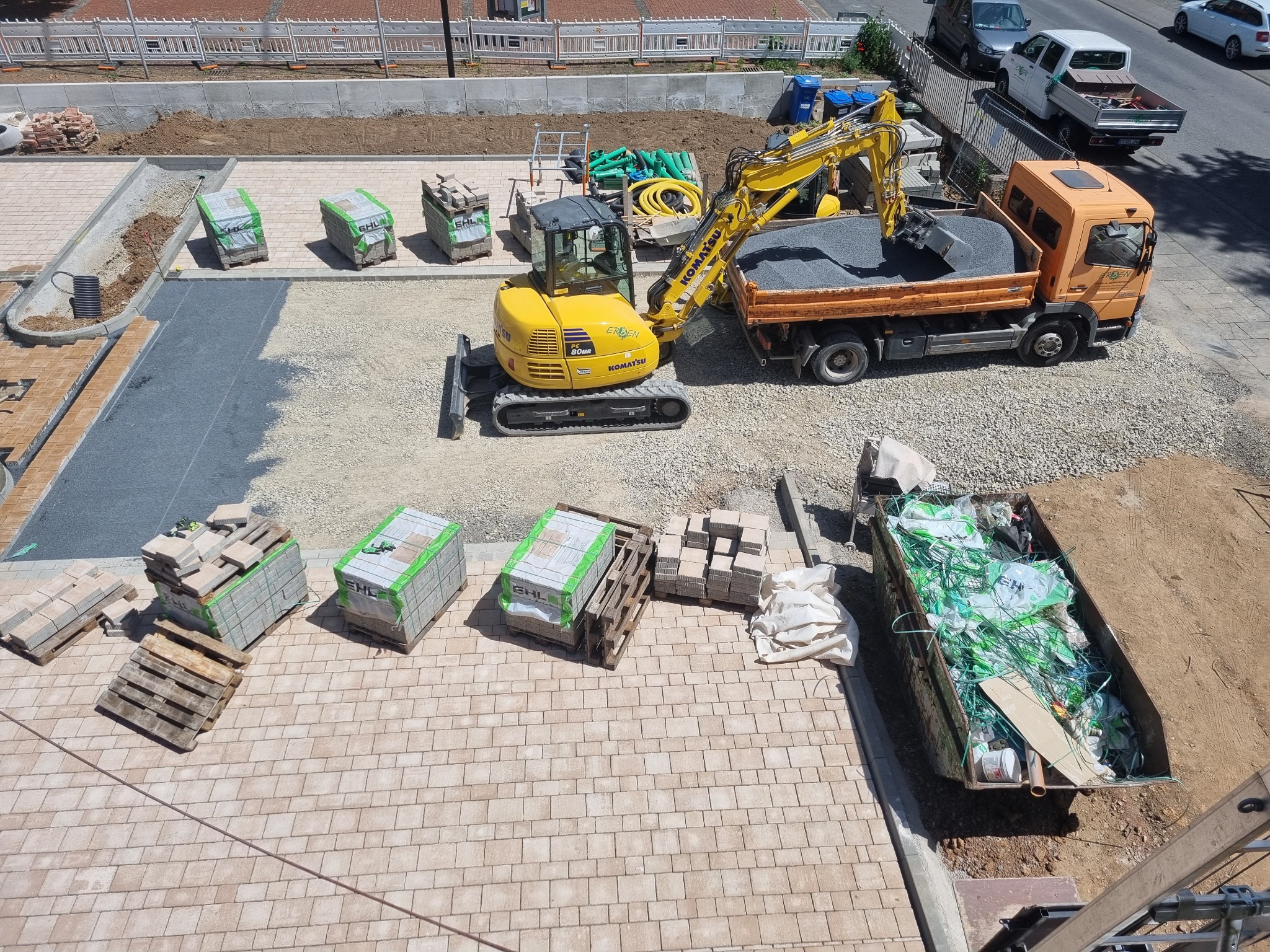 Widok z góry na plac budowy z żółtą koparką Komatsu ładującą żwir do pomarańczowej ciężarówki, palety z kostką brukową EHL, kontener na odpady wypełniony folią i gruzem oraz częściowo ułożona...