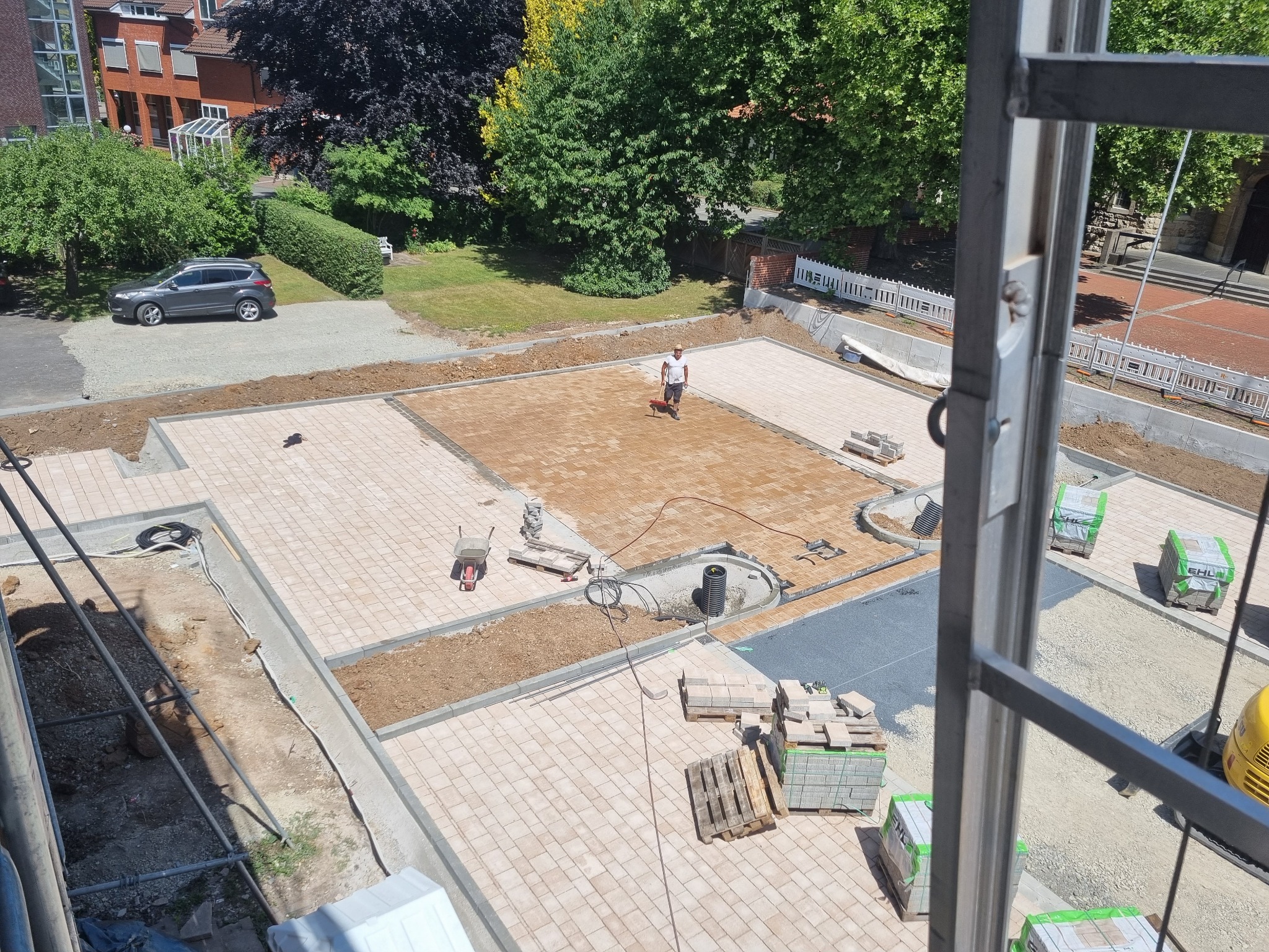 Widok z góry na plac budowy z częściowo ułożoną kostką brukową w odcieniach beżu i brązu, pracownik wyrównuje piasek pod dalsze układanie, palety z kostką czekają na ułożenie, w tle budynki i zieleń.