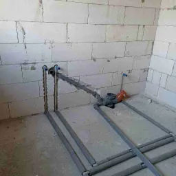 Surowe wnętrze z bloczków, widoczne szare rury instalacji wodnej lub grzewczej w trakcie montażu, niektóre z izolacją, z elementami armatury i podłączeniami do ściany.