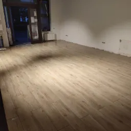 Świeżo ułożone, błyszczące panele podłogowe imitujące drewno w pustym pomieszczeniu z widocznymi niedokończonymi fragmentami ścian przy podłodze i otwartymi drzwiami wejściowymi.