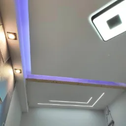 Nowoczesne oświetlenie sufitowe z liniowymi lampami LED tworzącymi geometryczny wzór, dodatkowo podświetlenie LED w kolorze fioletowym oraz lampy punktowe, widoczny fragment ściany z dekoracyjną...